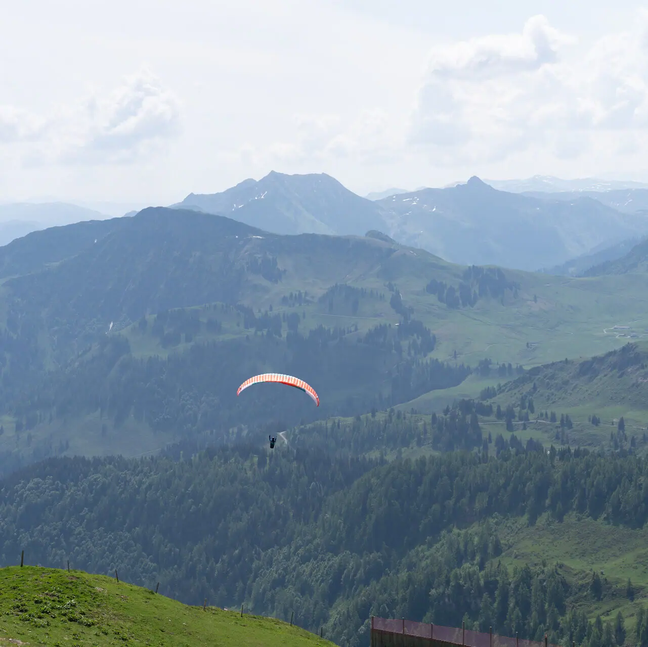 Eine Person beim Paragliding über einem Tal mit Bergen und Wolken im Hintergrund.