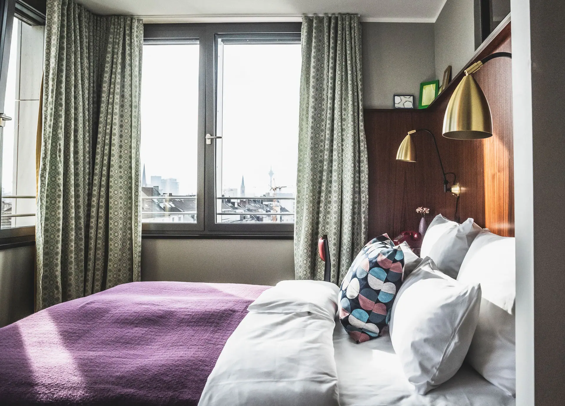 HENRI Düsseldorf Ein Bett mit Kissen und einer Lampe in einem Raum mit Fenstern.