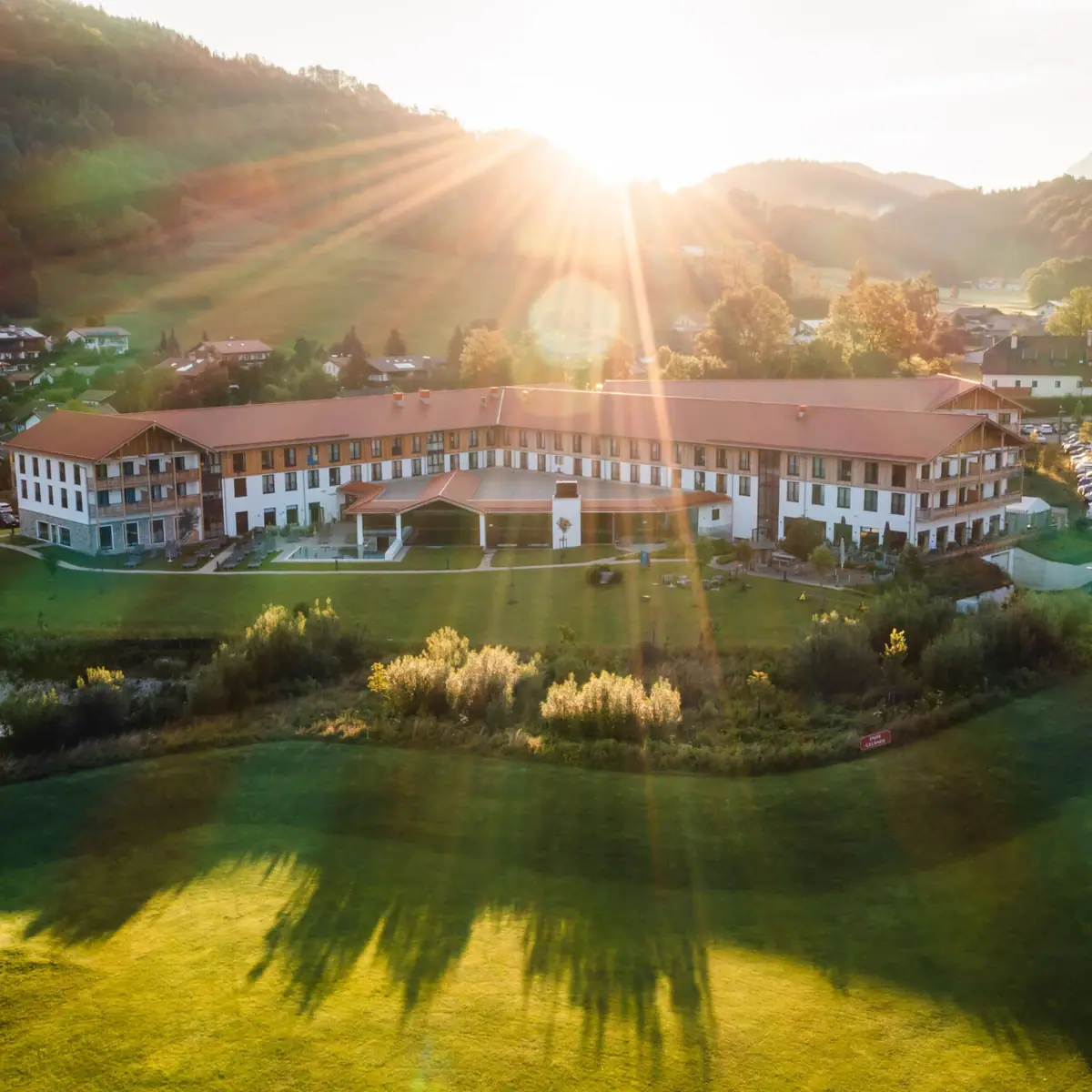 aja Hotel in Ruhpolding Luftaufnahme eines großen Hotels mit weitläufigem Garten in der Alpenlandschaft, Sonnenstrahlen strahlen über die Hügel und den Morgenhimmel