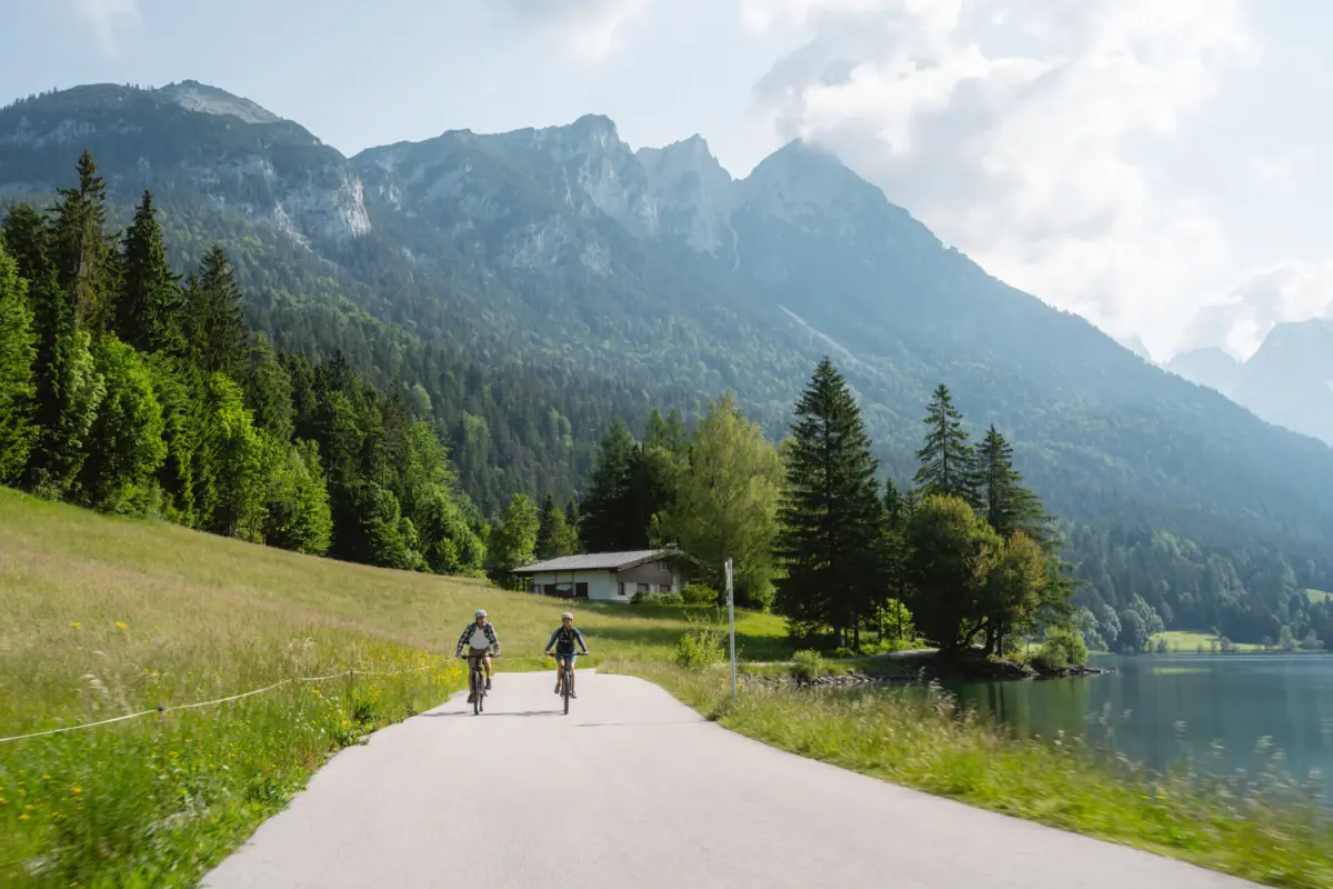 Radtour mit Bergen im Hintergrund Zwei Personen fahren auf Fahrrädern auf einer Straße in der Nähe eines Sees.