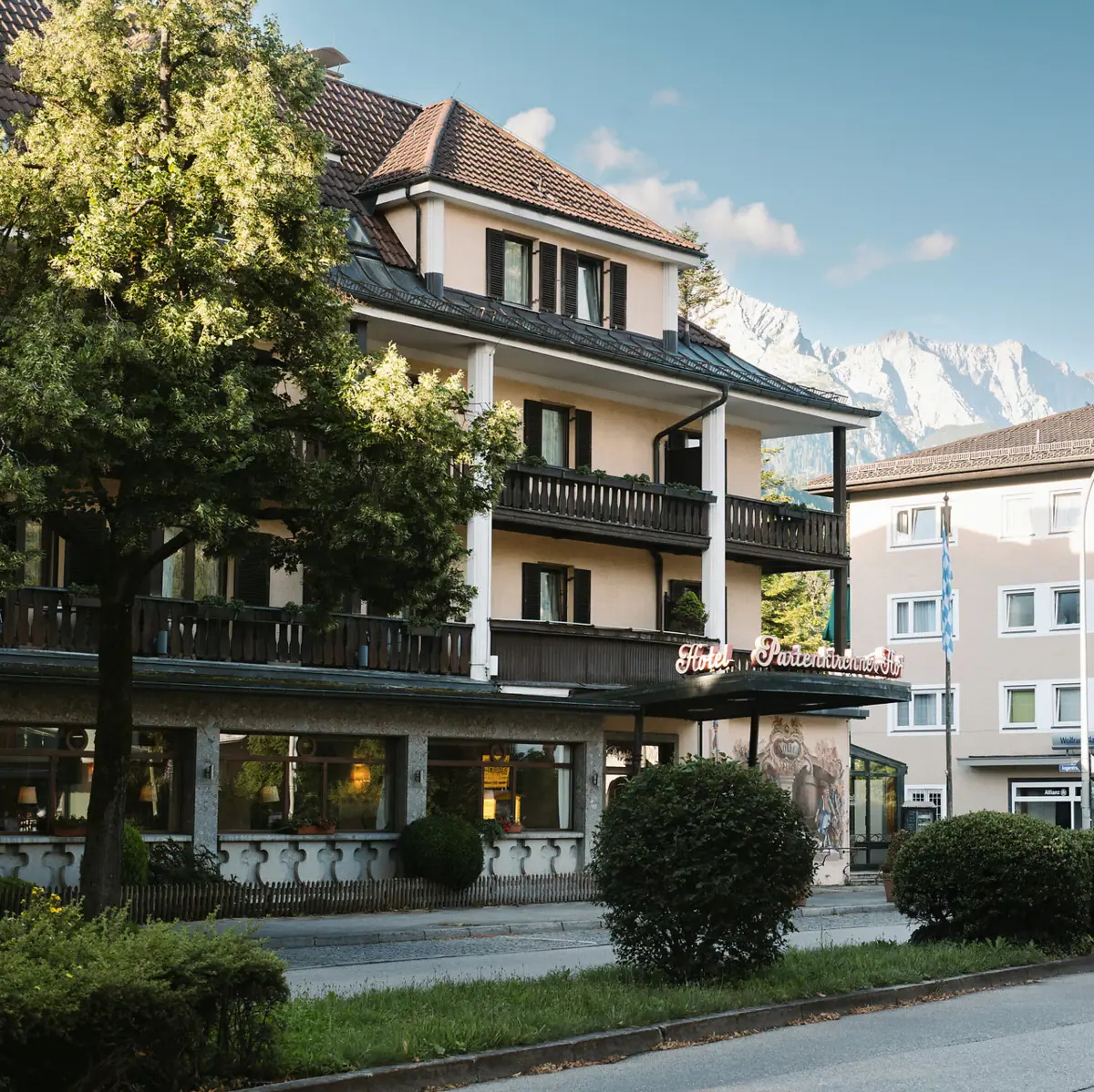 HENRI Hotel Garmisch-Partenkirchen Das HENRI Hotel in Garmisch-Partenlkirchen mit Bäumen am Straßenrand und Bergen im Hintergrund.