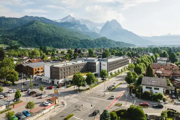 aja Garmisch-Partenkirchen Das aja Garmisch-Partenkirchen mit vielen Gebäuden und Bäumen unter einem bewölkten Himmel sowie Bergen im Hintergrund.