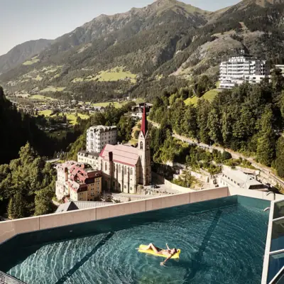 Rooftop Infinity-Pool Bad Gastein Eine Person schwimmt mit einer gelben Luftmatratze in einem Rooftop-Infinity-Pool auf einem hohen Gebäude mit Bergen im Hintergrund.