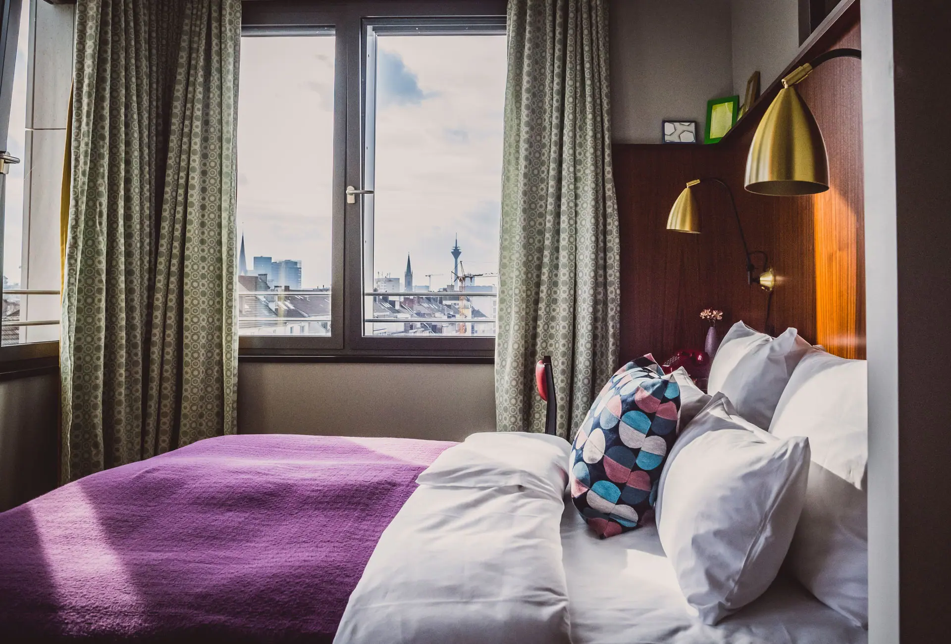 HENRI Hotel Düsseldorf Bett mit Kissen und lila Decke in einem Zimmer mit Fenster.