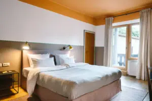 Hotelzimmer im HENRI Hotel Garmisch-Partenkirchen Ein gemütliches Bett in einem modernen Hotelzimmer im HENRI Hotel Garmisch-Partenkirchen mit einem Zugang zum Balkon im Hintergrund.