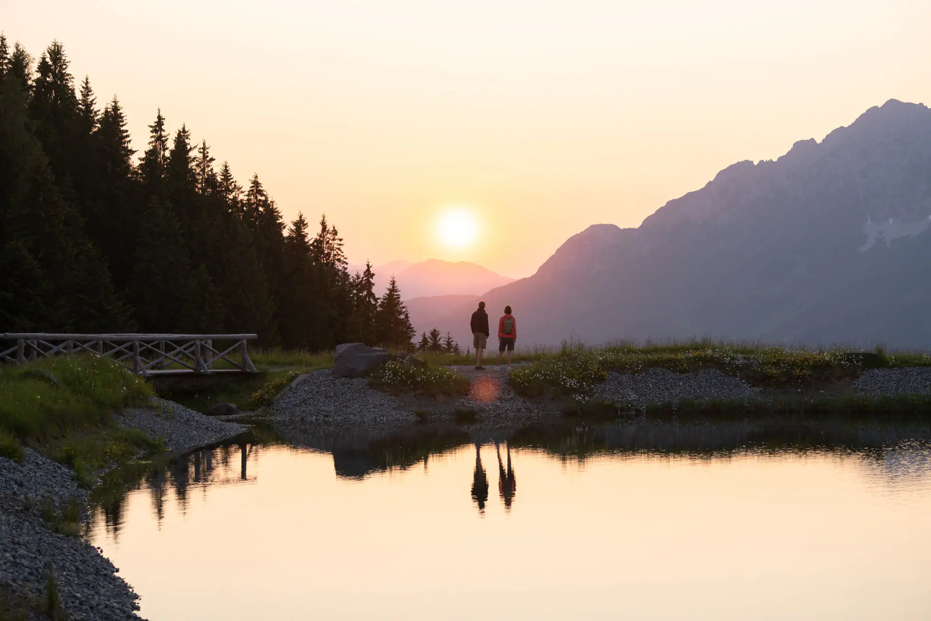 Bergsee in Kitzbühel Zwei Personen gehen an einem felsigen Ufer eines Sees entlang, mit Bäumen und Bergen im Hintergrund.